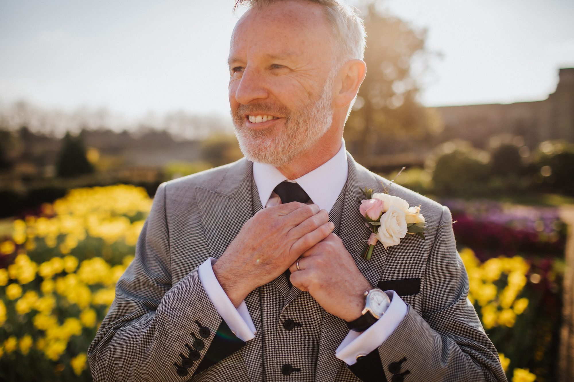 A groom in a grey three-piece wedding suit with a rose buttonhole adjusting his tie in a sunlit garden