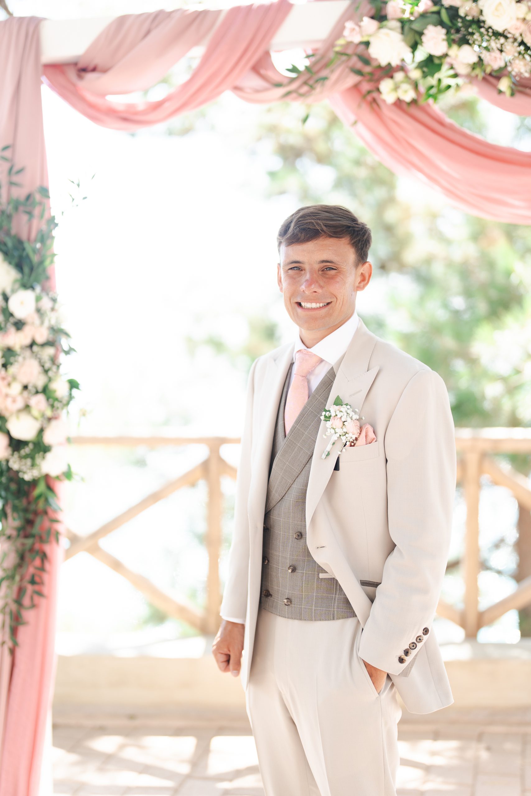 A groom in an ivory three-piece wedding suit standing beneath a floral wedding arch