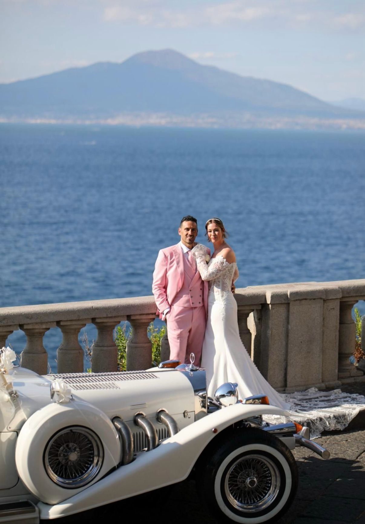 A bride and groom with a vintage white wedding car overlooking the sea